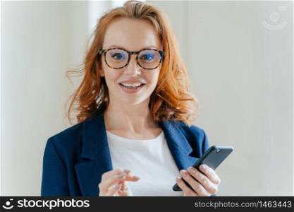 Headshot of female banker with pleasant appearance and red hair, does banking online on cell phone, checks notification on financial website, wears transparent glasses and elegant formal suit