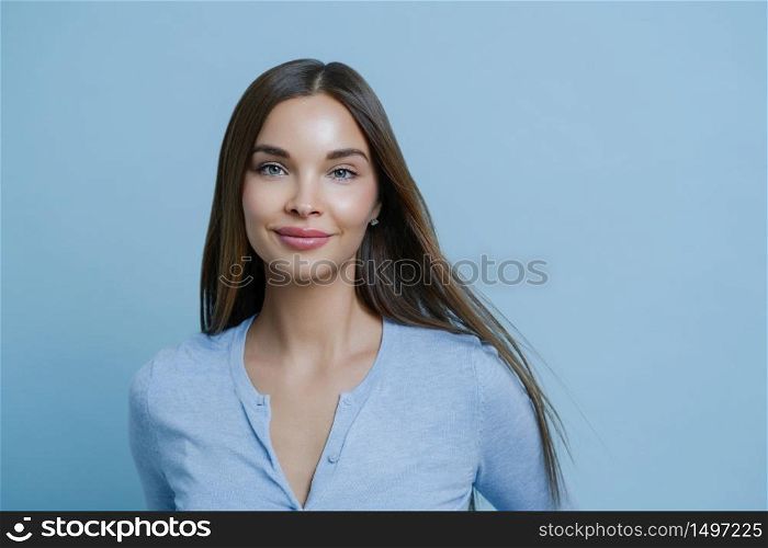 Headshot of good looking young woman with dark long hair, has tender feminine look, stands satisfied, wears blue jumper in one tone with studio background. People, lifestyle, skin care concept