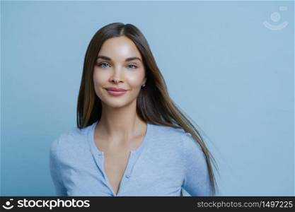 Headshot of good looking young woman with dark long hair, has tender feminine look, stands satisfied, wears blue jumper in one tone with studio background. People, lifestyle, skin care concept