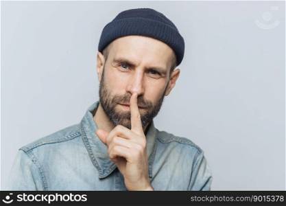 Headshot of handsome man with thick beard and mustache, has secret look, demonstrates silence sign, asks to be silent, isolated over grey background. Facial expressions and body language concept