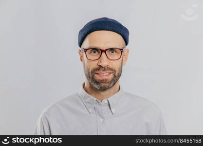 Headshot of handsome unshaven man wears transparent glasses, headgear, formal shirt, looks directly at camera, isolated over white background. Caucasian male with stubble dressed in fashionable outfit