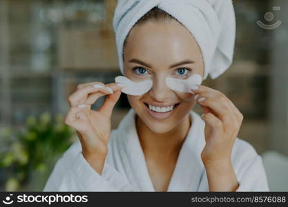 Headshot of happy young blue eyed woman with minimal makeup takes off beauty patches enjoys good effect has healthy well cared skin white perfect teeth wears bathrobe and wrapped towel on head