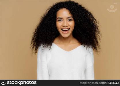 Headshot of healthy positive female model laughs happily, has bushy Afro hair, dressed in white soft jumper, glad to receive compliment from boyfriend, isolated over brown background. Emotions concept