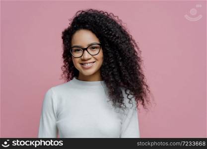 Headshot of lovely curly haired woman with healthy skin and toothy smile, dressed in white jumper, wears transparent glasses, isolated over rosy background. People, face expressions and emotions