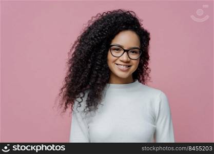Headshot of lovely curly haired woman with healthy skin and toothy smile, dressed in white jumper, wears transparent glasses, isolated over rosy background. People, face expressions and emotions