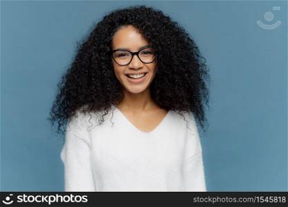 Headshot of lovely curly woman being in good mood, smiles broadly, expresses nice feelings, wears transparent spectacles and white jumper, isolated over blue background. Afro female poses indoor