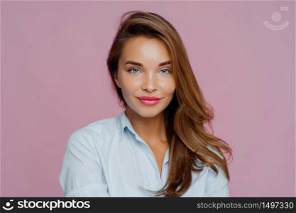 Headshot of lovely European lady has blue eyes, red painted lips, healthy skin, long hair, looks directly at camera, wears shirt, poses over purple background. Beautiful woman ready for job interview