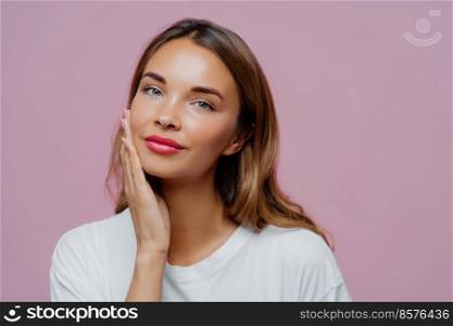 Headshot of lovely female model touches gently cheek, enjoys delicate face skin, wears makeup, has manicure, cares about her beauty, looks straightly at camera, dressed in white jumper, models indoor