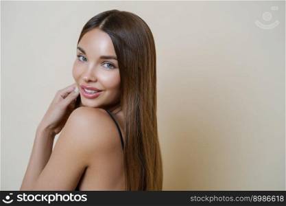 Headshot of lovely female model with glowing healthy skin, gentle smile, has long straight hair, looks with positive expression at camera, models against beige background, copy space. Beauty portrait