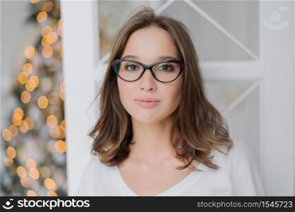 Headshot of lovely young female model in transparent glasses, has dark hair, dressed in casual clothes, enjoys domestic atmosphere, poses against New Year tree. People and lifestyle concept.