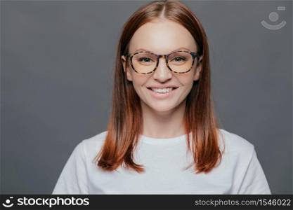 Headshot of pleasant looking dark haired young woman wears transparent glasses, dressed in casual clothes, has healthy skin, positive look, shows white perfect teeth, isolated over grey background.