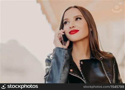 Headshot of pleasant looking dreamy woman focused aside, has telephone conversation, wears leather coat, notices someone into distance, poses over blurred background. Good roaming connection