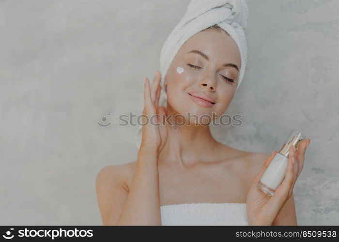 Headshot of pleased attractive woman applies face lotion, satisfied with new cosmetic product, keeps eyes closed, touches soft skin after bath, has well cared complexion, poses against grey wall