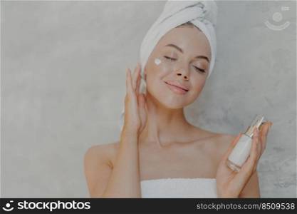 Headshot of pleased attractive woman applies face lotion, satisfied with new cosmetic product, keeps eyes closed, touches soft skin after bath, has well cared complexion, poses against grey wall
