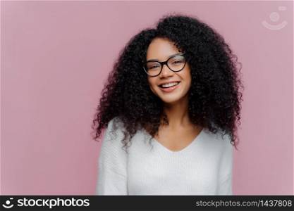 Headshot of pleased lovely woman with curly hairstyle, smiles gently at camera, wears optical glasses and white casual sweater, poses against purple background. Positive emotions and feelings concept