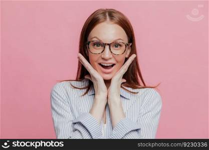 Headshot of positive beautiful business lady keeps both hands under chin, dressed in elegant shirt, looks with happiness at camera, opens mouth, isolated over pink background. Emotions concept