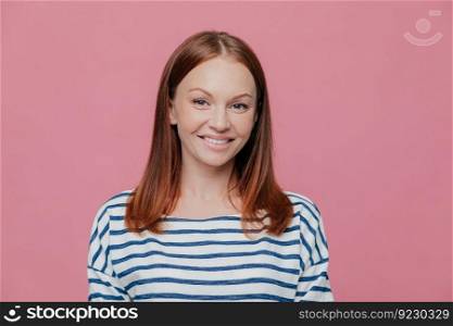 Headshot of pretty smiling European woman with charming smile, wears striped jumper, has brown hair, looks directly at camera, isolated over pink studio background. People, joy, happiness concept