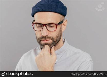 Headshot of sad lonely serious male keeps hand under chin, has dark bristle, looks down, thinks about promotion and starting new career, wears spectacles, hat and shirt, isolated over grey studio wall