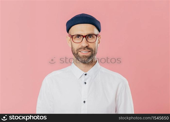 Headshot of satisfied bearded young man looks confidently at camera through spectacles, wears white formal shirt, blue hat, models against pink background, prepares for meeting with investors