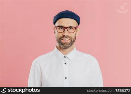 Headshot of satisfied bearded young man looks confidently at camera through spectacles, wears white formal shirt, blue hat, models against pink background, prepares for meeting with investors