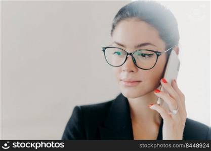 Headshot of serious brunette female manager with makeup wears trendy eyeglasses, formal clothes, has red manicure, calls business partner during remote job, poses indoor against white background