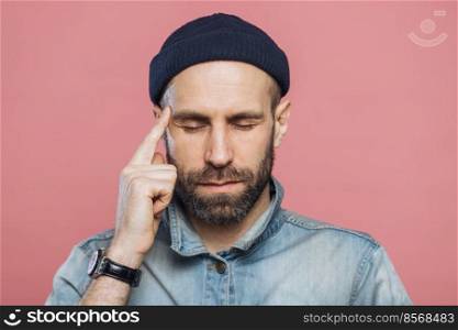 Headshot of serious thoughtful middle aged man tries to remember something in his mind, keeps fore finger on temple, keeps eyes shut, isolated over pink background, wears fashionable clothing