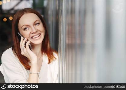 Headshot of smiling female employee with cheerful facial expression, uses smart phone gadget for discussion information with friend, makes mobile conversation, wears formal clothes, stands indoor