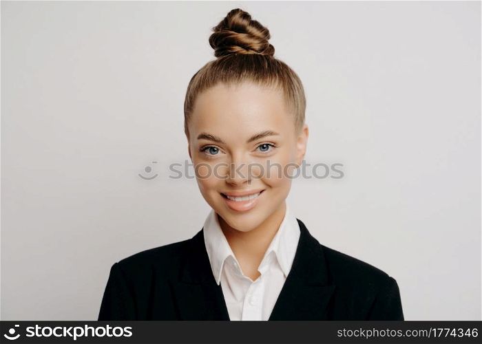 Headshot of smiling happy attractive business woman in dark suit with hair in bun standing isolated in front of spacious grey background while looking straight forward with calm and asureness. Smiling attractive business woman in dark suit