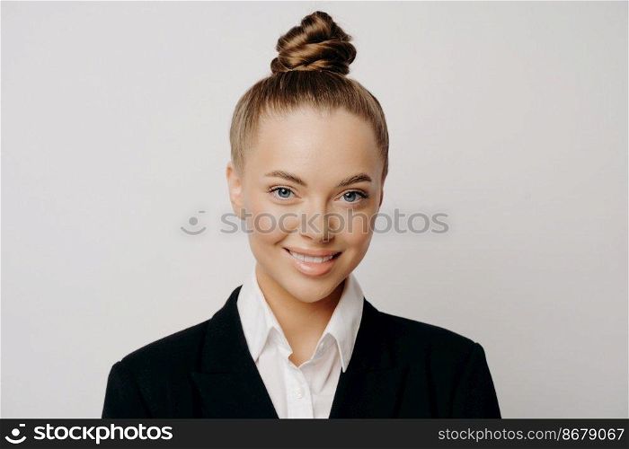 Headshot of smiling happy attractive business woman in dark suit with hair in bun standing isolated in front of spacious grey background while looking straight forward with calm and asureness. Smiling attractive business woman in dark suit