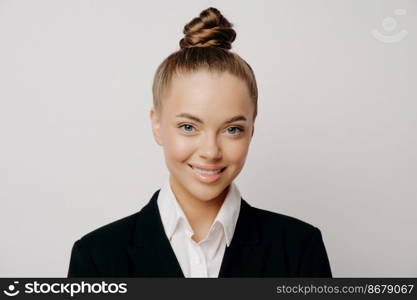 Headshot of smiling happy attractive business woman in dark suit with hair in bun standing isolated in front of spacious grey background while looking straight forward with calm and asureness. Smiling attractive business woman in dark suit