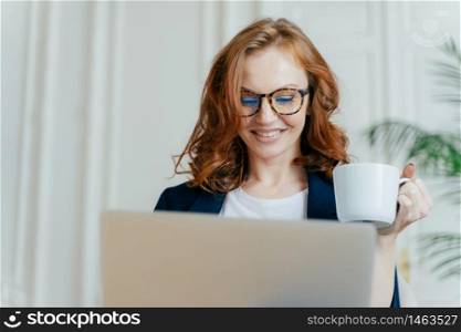 Headshot of smiling red haired female in optical glasses, enjoys hot coffee or tea, focused in laptop computer, makes purchases online, smiles pleasantly, works freelance on distance, books tickets
