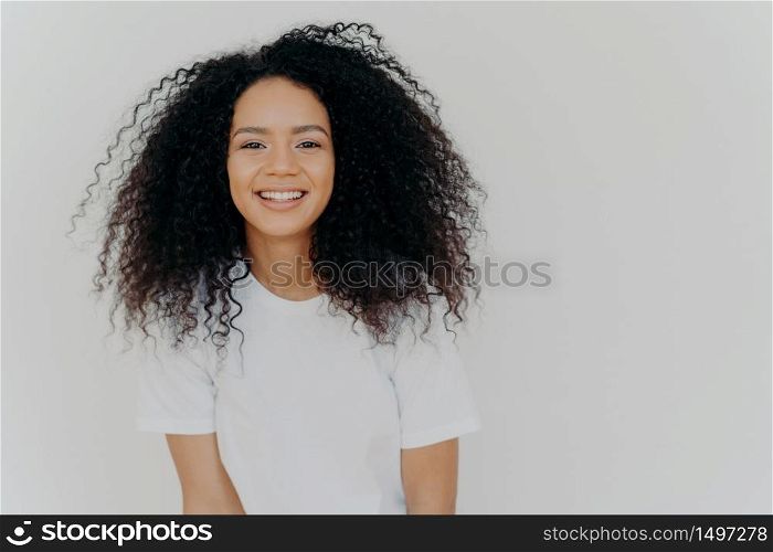 Headshot of smiling woman has bushy luminous hair, stands confident and pleased, minimal makeup, enjoys lively small talk, dressed in comfortable white t shirt, poses indoor. Happy emotions.