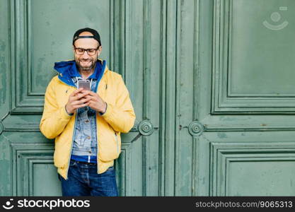 Headshot of stylish man with beard and broad forehead in yellow anorak, jeans and cap in big glasses holding cell phone checking newsfeed on social network accounts isolated over green ancient door