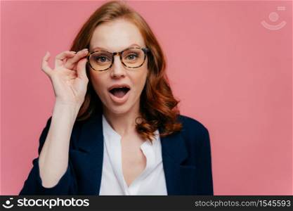Headshot of surprised red haired woman keeps hand on frame of glasses, jaw dropped, can not believe her eyes, wears elegant suit, shocked by unexpected rumor, stands over pink studio wall, free space