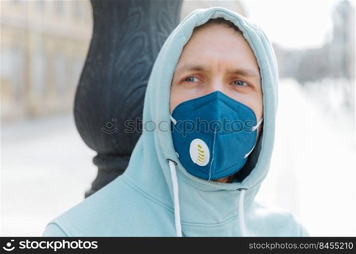 Headshot of thoughtful young man wears hood and respiratory mask, walks on street, protects from coronavirus, thinks about dangerous disease, being in danger. Spreding Covid-19 around world.