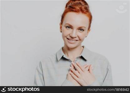 Headshot of young grateful ginger holding hands on chest, feeling love and appreciation, lovely red haired caucasian female thanking while posing isolated on grey studio background. Body language. Headshot of young grateful ginger holding hands on chest, feeling love and appreciation