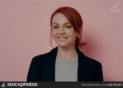Headshot portrait of charming young red-haired female smiling pleasantly at camera and feeling happy after working day in office, wearing jacket over basic t-shirt, isolated on rosy background. Charming young red-haired female smiling pleasantly at camera, isolated on rosy background