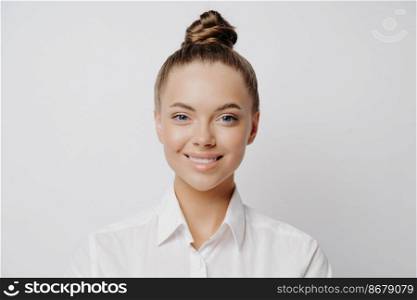 Headshot portrait of happy cheerful female worker with bright smile in white shirt feeling excited to hear about further developments at her workplace, standing alone in front of light wall. Smiling official female worker in white shirt