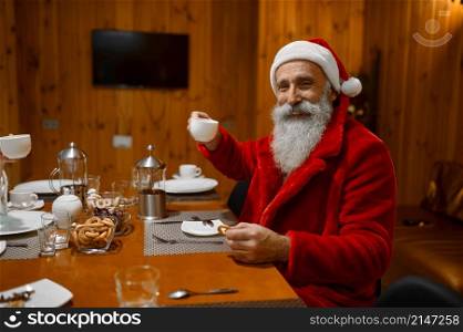 Headshot portrait of happy smiling Santa Claus drinking tea and eating bagels in sauna rest room. Portrait of happy smiling Santa drinking tea