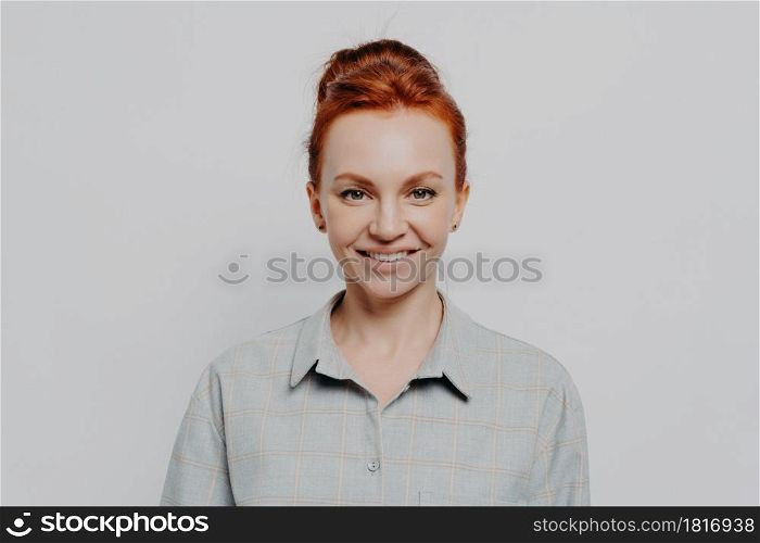 Headshot portrait of happy young caucasian ginger woman being satisfied and feeling happy, looking at camera cheerful while standing isolated on grey background. Positive emotions concept. Headshot portrait of happy young caucasian ginger woman being satisfied and feeling happy