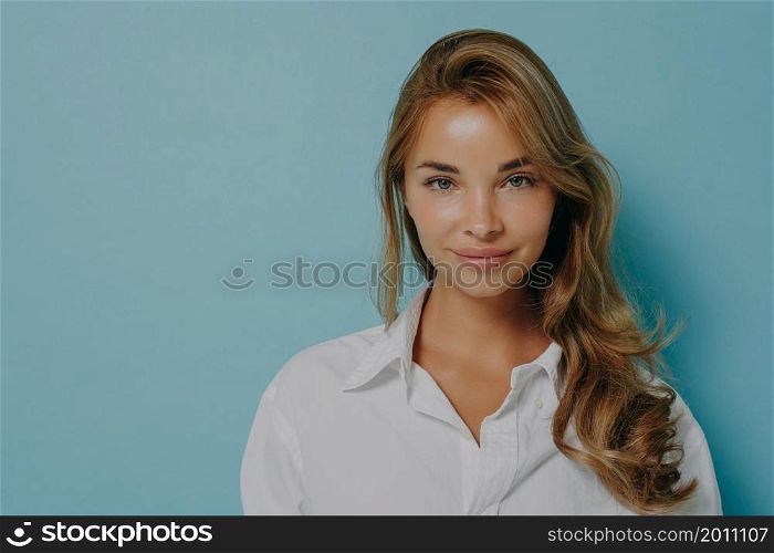 Headshot portrait of young caucasian female model with dyed blonde hair in casual white shirt posing with pleasant smile isolated over light blue studio wall background with copy space for advertising. Portrait of young caucasian female model with dyed blonde hair smiling at camera on blue background