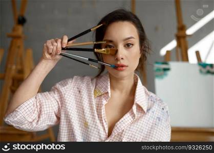 Headshot portrait of young woman artist holding paintbrush near her face posing over art studio workshop. Headshot portrait of young woman artist holding paintbrush near her face