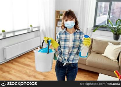 health and hygiene concept - smiling asian woman wearing protective medical mask for protection from virus holding bucket of cleaning stuff and detergent at home. asian woman in protective mask cleaning home