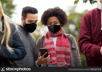 health and people concept - group of friends with smartphone wearing face protective medical mask for protection from virus disease outdoors. friends in masks with smartphone outdoors