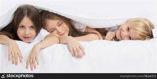 Health, beauty and childhood concept - three smiling children waking up in bed under one blanket at home. Children waking up in bed