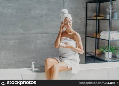 Health, beauty and p&ering concept. Beautiful dreamy young Eurpean woman looks away thoughtfully, applies body cream, sits in bathroom, wrapped in white soft towel, uses nice cosmetic product