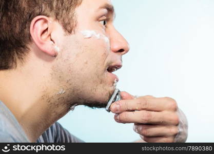 Health beauty and skin care concept. Closeup young bearded man with foam on face shaving with razor on blue background.