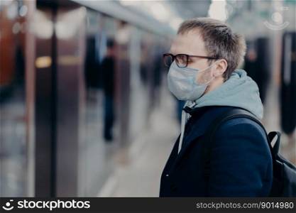 Health care and illness concept. Profile shot of European man wears medical mask, poses on station in underground, takes preventive measures during quarantine, afraids of catching serious virus