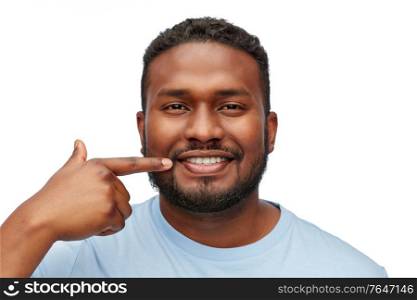health care, dental hygiene and people concept - portrait of happy smiling young african american man pointing finger to his mouth or teeth over white background. smiling man pointing finger to his mouth or teeth