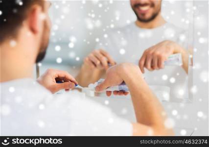 health care, dental hygiene, people and beauty concept - close up of young man cleaning teeth and squeezing toothpaste on toothbrush at home bathroom over snow